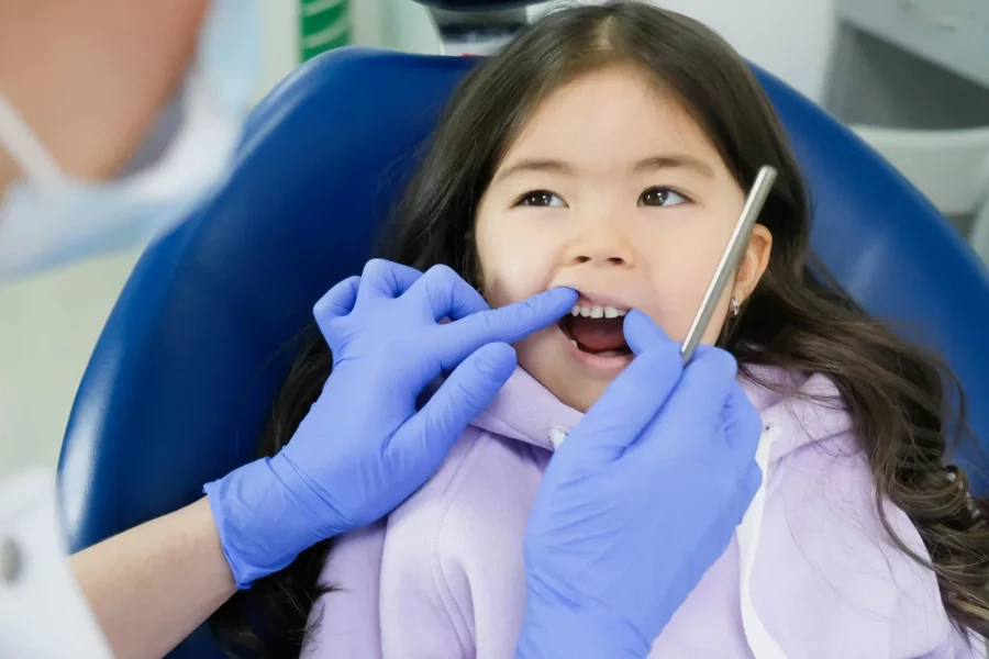 Kind auf Zahnarztstuhl mit lächelndem Gesicht, Zahnarzt hält sanft die Lippe hoch, weiße Milchzähne sichtbar.